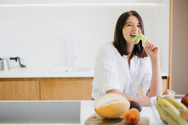 Young happy woman eating green lettuce leaf and smiling on background of fresh fruits and vegetables in modern white kitchen. Healthy food concept. Home cooking